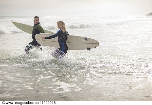 Paar mit Surfbrettern  die zusammen durch die Wellen laufen.