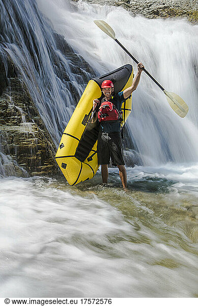 Paddler stands beside waterfall  raising paddle in the air.