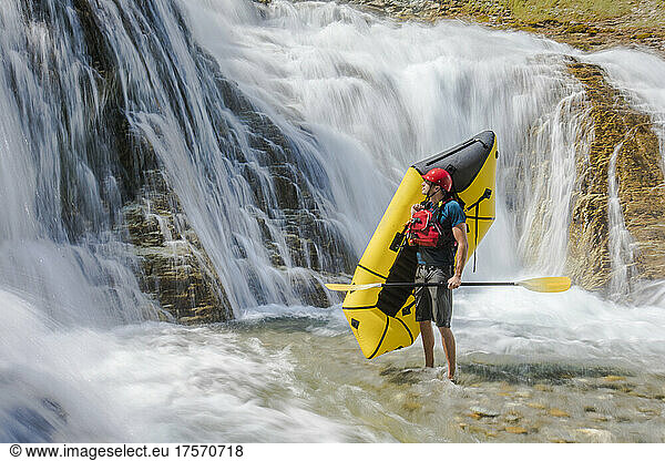 Paddler stands holding raft (packraft) below large waterfall.