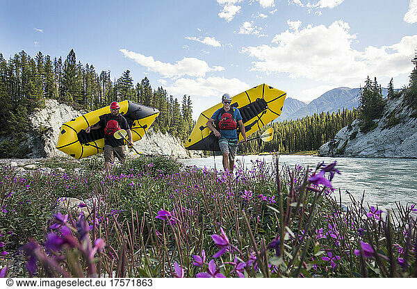 Paddlers carry yellow packrafts beside scenic river  flowers