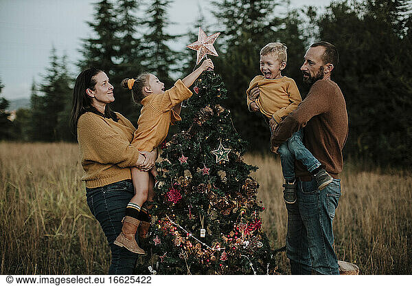 Parents carrying happy children decorating Christmas tree at countryside during sunset
