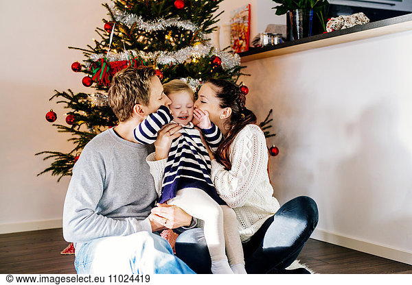 Parents kissing daughter while sitting against Christmas tree at home
