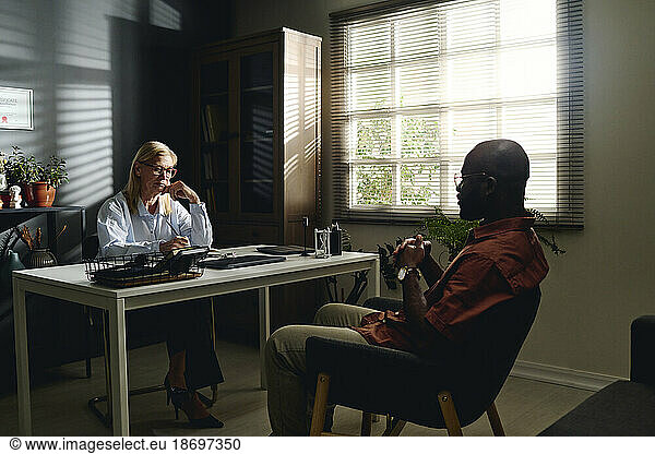 Patient sitting on chair with psychologist near desk at office