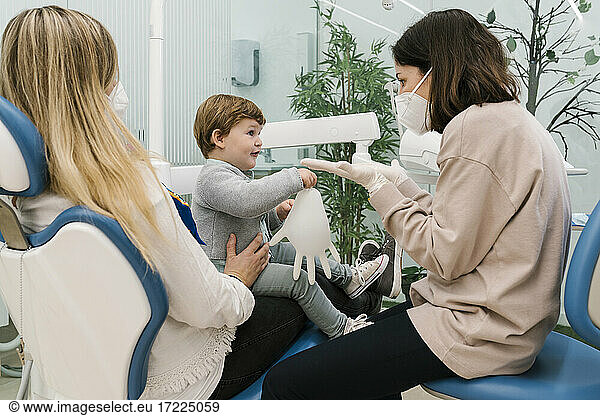 Pediatric dentist wearing protective face mask talking to boy sitting with mother in clinic