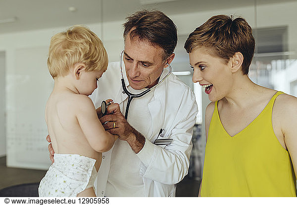 Pediatrician examining toddler with stethoscope  mother standing next to them