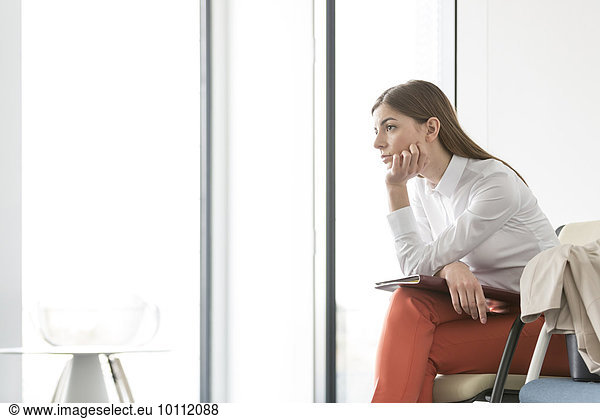 Pensive brunette businesswoman looking out office window with hand on chin