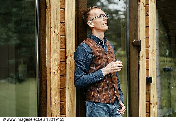 Pensive middle age man stands on the terrace of a cozy bungalow and drinks bourbon