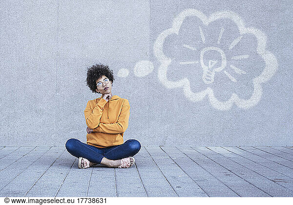 Pensive woman sitting on ground with electric bulb in thhought bubble