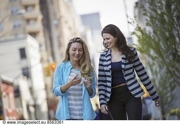 People outdoors in New York city in spring time. Two women walking  one checking her cell phone.