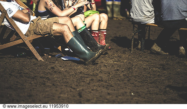 People sitting on deckchairs in the mud  wearing Wellington boots  at a summer music festival.