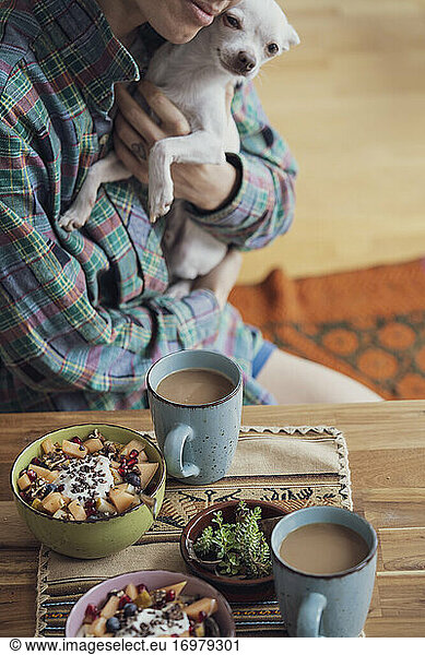 person cuddles cute puppy dog at table with healthy breakfast & coffee