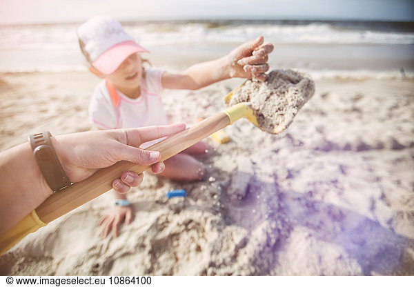 Personal perspective of mother on beach holding shovel of sand for daughter