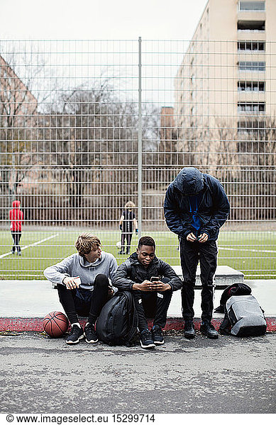 Phone addicted friends on sidewalk against playing field in city