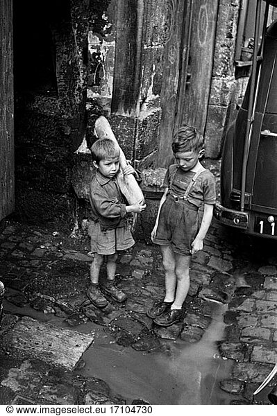 Photography:
children’s photographs. Street scene: two little boys look at a stream of water running at their feet down the pavement. One of the boys carries a large loaf of bread. Photo  Rouen (France)  undated.