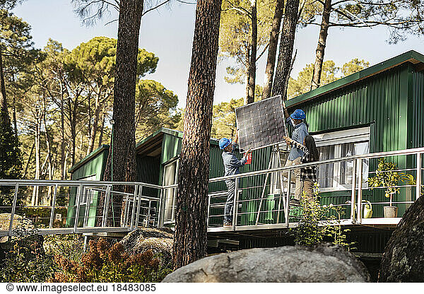 Photovoltaic technicians mounting solar panel on sustainable house with home owner standing on veranda