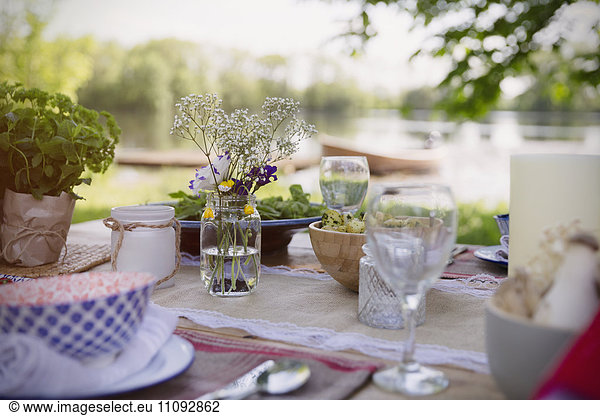 Platzieren Sie Einstellungen und einfachen Blumenstrauß auf dem Gartenparty-Tisch am Seeufer.