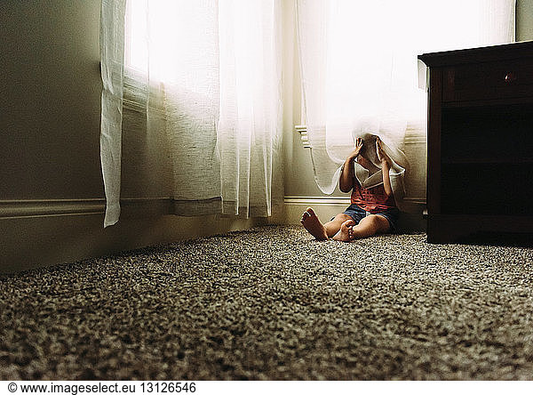 Playful boy hiding behind curtain while sitting on rug at home