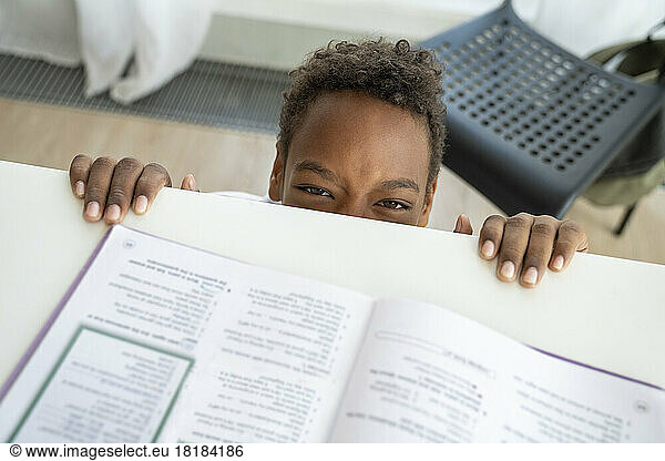 Playful boy peeking from study table at home