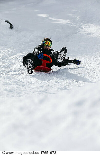 Playful boy wearing ski goggles tobogganing through snow