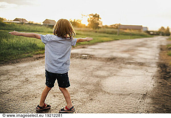 Playful boy with arms outstretched standing on road at sunset