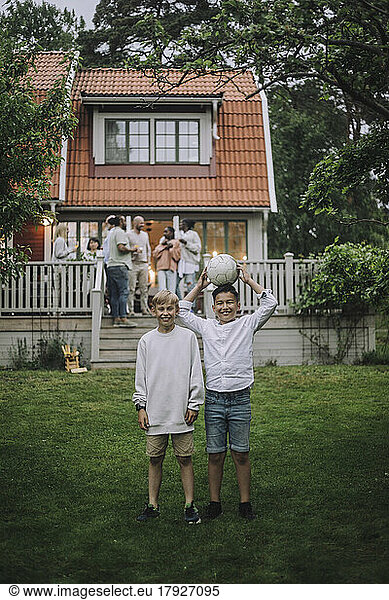 Playful boys with soccer ball standing in back yard with family on porch in background