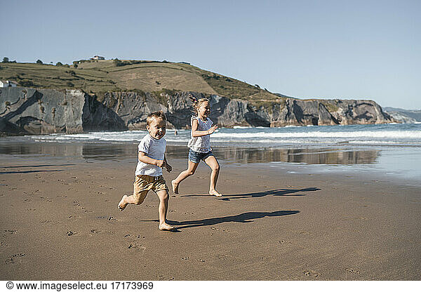 Playful brother and sister running at beach against hill during sunny day