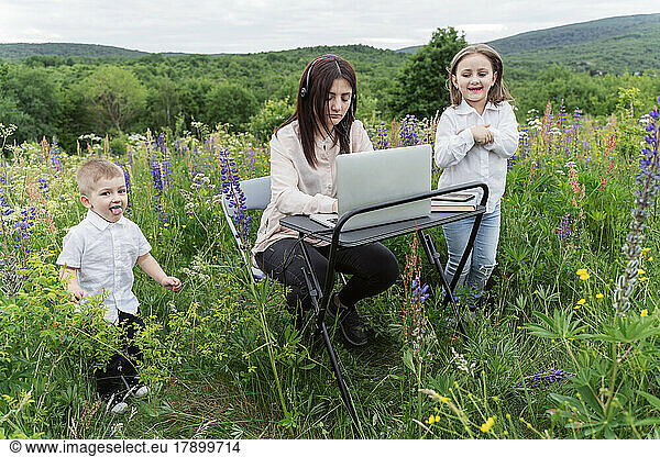 Playful children by mother working on laptop in meadow
