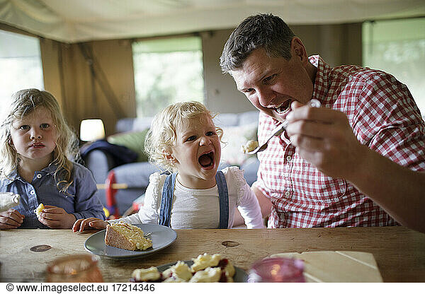 Playful father feeding cake to cute eager daughter