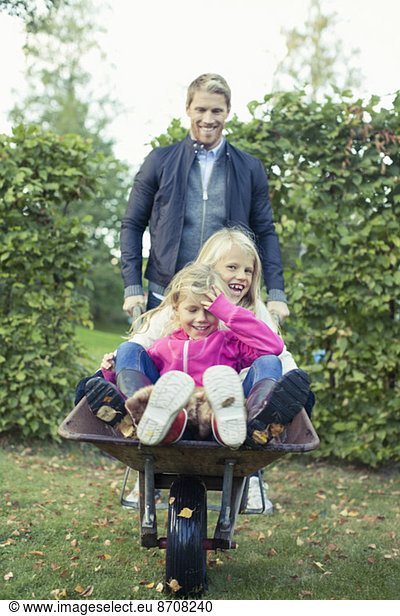 Playful father pushing daughters on wheelbarrow at yard