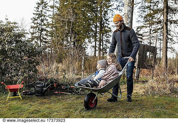 Playful father wheeling son and daughter sitting in wheelbarrow at back yard