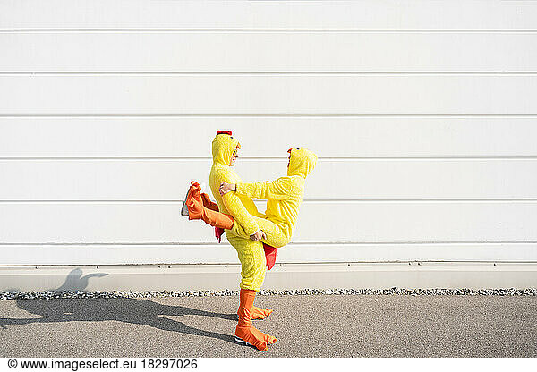 Playful friends wearing chicken costumes standing by white wall on sunny day