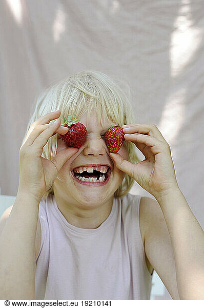 Playful girl covering eyes with fresh strawberries from garden