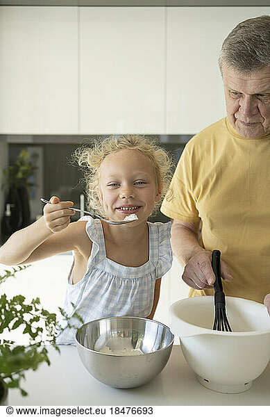 Playful girl holding spoon by grandfather preparing batter in kitchen