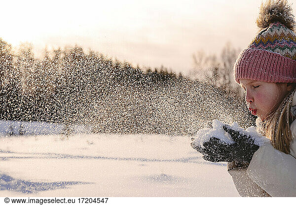 Playful girl in warm clothing blowing snow during winter