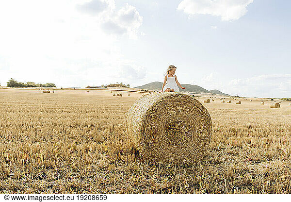 Playful girl sitting on bale of straw in stubble field on sunny day