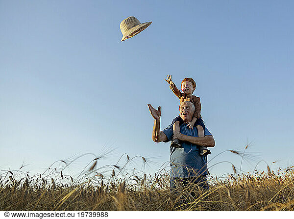 Playful grandfather carrying grandson on shoulders and throwing straw hat under sky