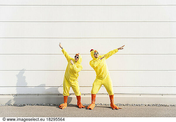 Playful man and woman wearing chicken costumes gesturing and having fun in front of wall