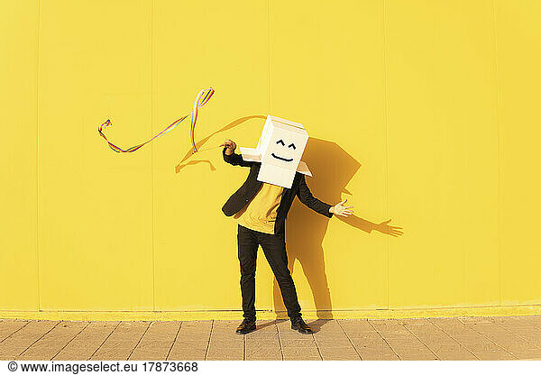 Playful man wearing box with smiley face spinning rainbow colored rhythmic gymnastics ribbon in front of yellow wall