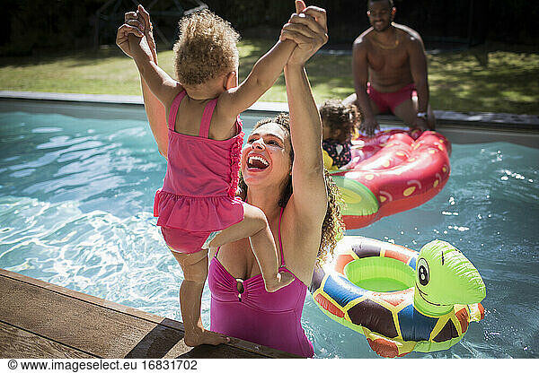 Playful mother lifting toddler daughter into sunny swimming pool