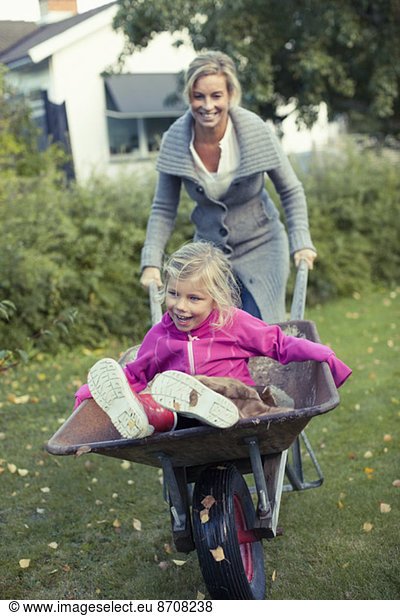Playful mother pushing daughter on wheelbarrow at yard