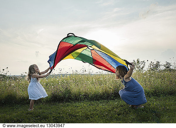 Playful sisters playing with colorful textile on grassy field against sky