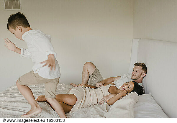 Playful son running on bed while parents lying in bedroom