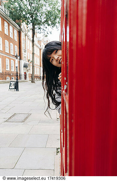 Playful woman peeking by telephone booth in city