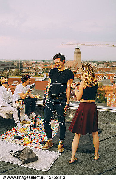 Playful woman tying string light around man while friends enjoying at rooftop party
