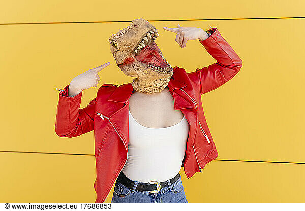 Playful woman wearing dinosaur mask pointing in front of yellow wall