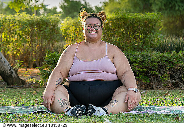 Plus sized woman sitting on yoga mat outdoors smiling while meditating