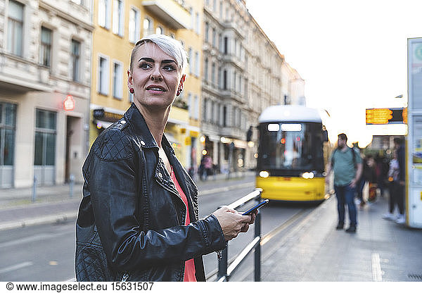 Porträt einer blonden Frau mit Smartphone an der Straßenbahnhaltestelle  Berlin  Deutschland
