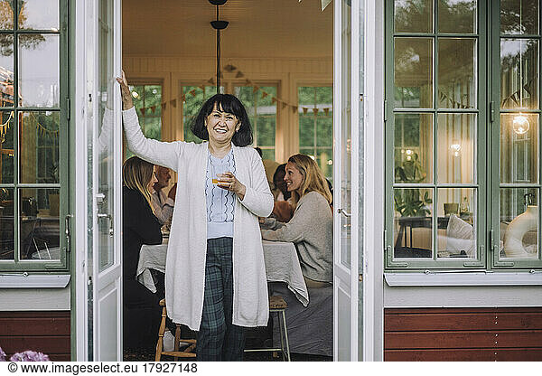 Porträt einer Frau mit Strickjacke  die ein Glas in der Hand hält und an der Tür steht