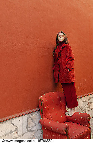 Porträt einer in Rot gekleideten jungen Frau  die barfuss auf der Armlehne eines roten Loungesessels steht und an einer roten Wand lehnt