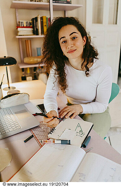 Porträt einer jungen Frau mit lockigem Haar  die am Schreibtisch Hausaufgaben macht